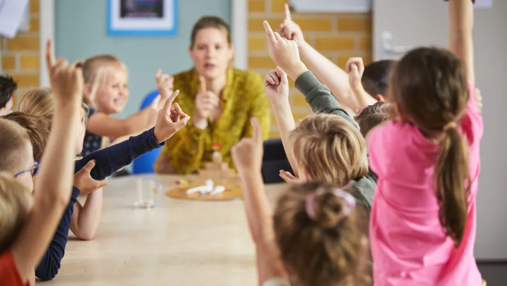 Foto van leerkracht voor klaslokaal met leerlingen die hun hand opsteken.