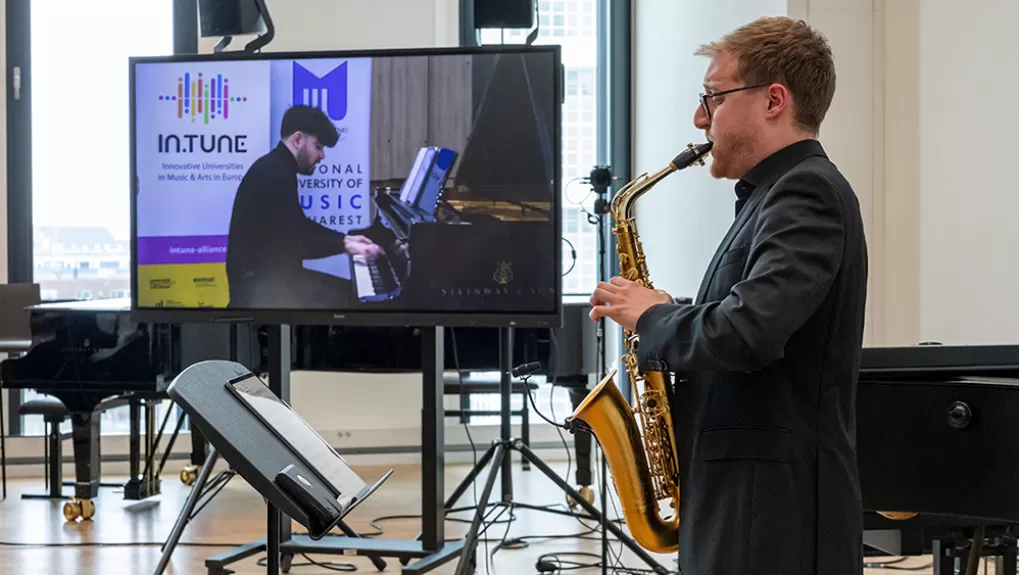 Een saxofonist op de voorgrond, met achter hem op een scherm een pianist. Ze spelen samen een duet, via digitale verbinding