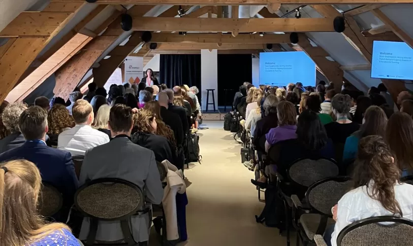 Foto van achteren tijdens een plenaire sessie tijdens de TCA. Je ziet Noor Groenendijk vooraan spreken, en kijkt bij de deelnemers op de rug. Ze zitten in rijen, met een pad in het midden als doorbreking.