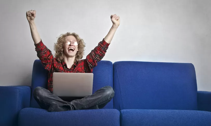 Man zit op bank, met laptop op schoot en juicht met zijn handen in de lucht, mond open