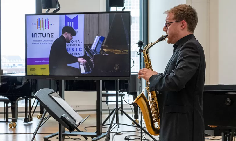 Een saxofonist op de voorgrond, met achter hem op een scherm een pianist. Ze spelen samen een duet, via digitale verbinding