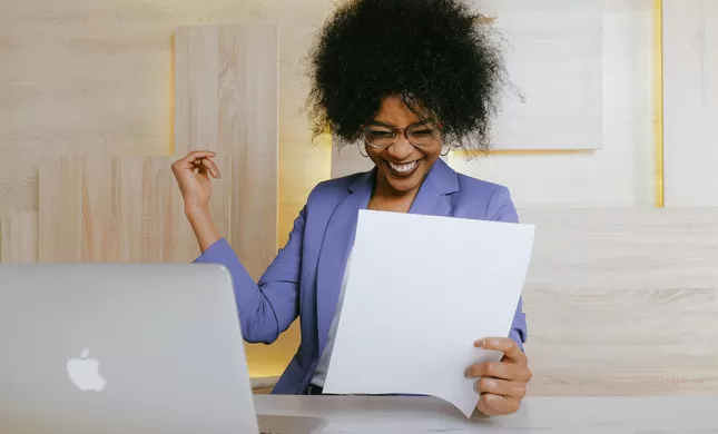 Vrouw achter laptop met papier in haar hand, ze kijkt heel erg blij