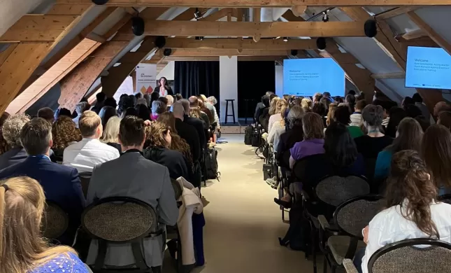Foto van achteren tijdens een plenaire sessie tijdens de TCA. Je ziet Noor Groenendijk vooraan spreken, en kijkt bij de deelnemers op de rug. Ze zitten in rijen, met een pad in het midden als doorbreking.