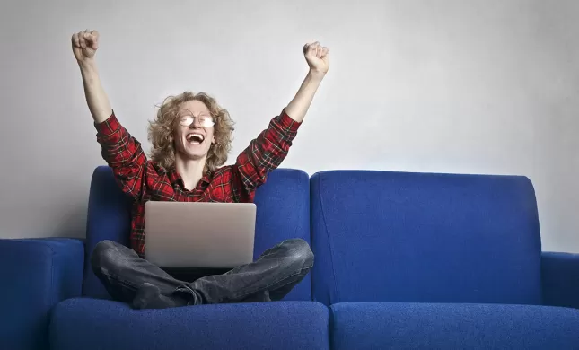 Man zit op bank, met laptop op schoot en juicht met zijn handen in de lucht, mond open
