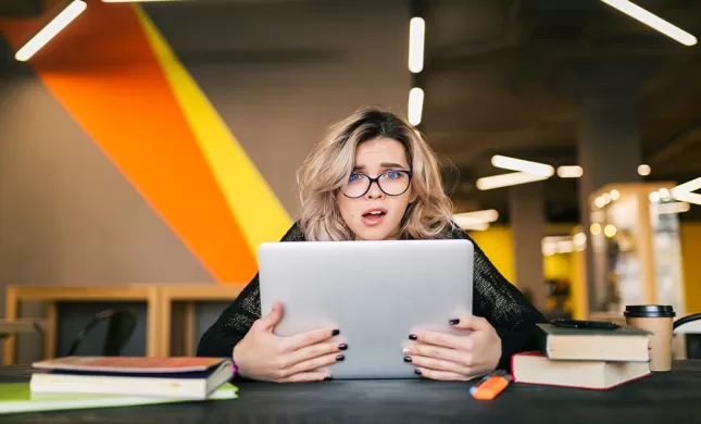Jonge vrouw zit in een bibliotheek-achtige ruimte achter haar laptop en pakt haar scherm vol ongeloof vast. Vrouw is blond, middellang haar, met een bril op.