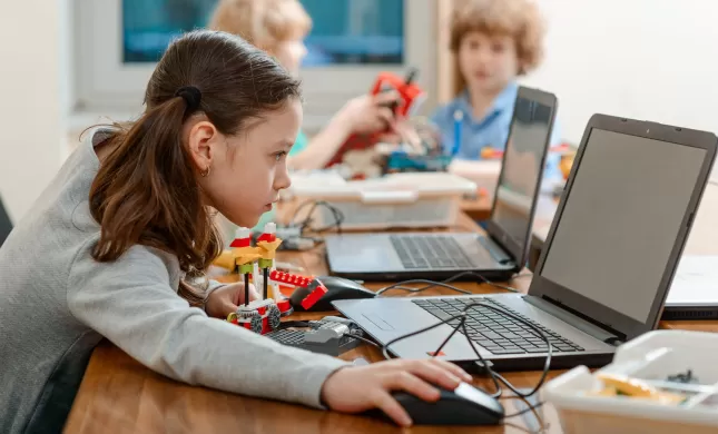 Meisje met twee staartjes achter een laptop. Ze is nog jong, zit op de basisschool en heeft ook een bouwsel voor zich dat gelinkt zit aan de laptop. Ze kijkt geconcentreerd naar het scherm. Achter haar zie je klasgenoten ook achter een laptop.