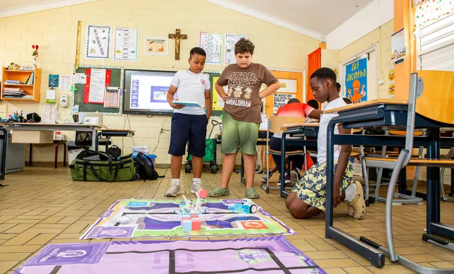 3 leerlingen in een klaslokaal op Curacao, staan en zitten om een mat heen, waarop een spel is afgebeeld. De linkertwee studenten staan, rechter zit op een stoel