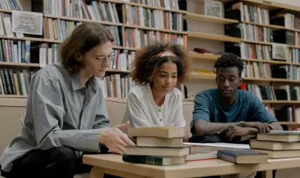 Drie studenten in een bibliotheek achter een tafel vol boeken. 2 mannen en in het midden een vrouw