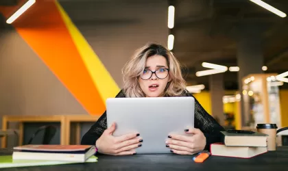 Jonge vrouw zit in een bibliotheek-achtige ruimte achter haar laptop en pakt haar scherm vol ongeloof vast. Vrouw is blond, middellang haar, met een bril op.