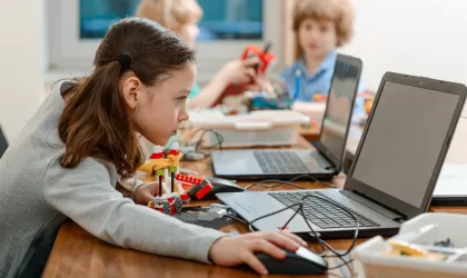 Meisje met twee staartjes achter een laptop. Ze is nog jong, zit op de basisschool en heeft ook een bouwsel voor zich dat gelinkt zit aan de laptop. Ze kijkt geconcentreerd naar het scherm. Achter haar zie je klasgenoten ook achter een laptop.