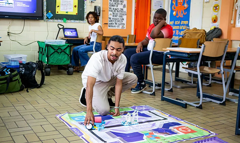Je ziet een speelmat op de grond van een leslokaal in Curacao. Op de achtergrond zitten leerlingen op een stoel achter hun eigen tafel. Op de voorgrond zit één student die met een object over de speelmat navigeert