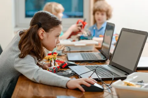 Meisje met twee staartjes achter een laptop. Ze is nog jong, zit op de basisschool en heeft ook een bouwsel voor zich dat gelinkt zit aan de laptop. Ze kijkt geconcentreerd naar het scherm. Achter haar zie je klasgenoten ook achter een laptop.
