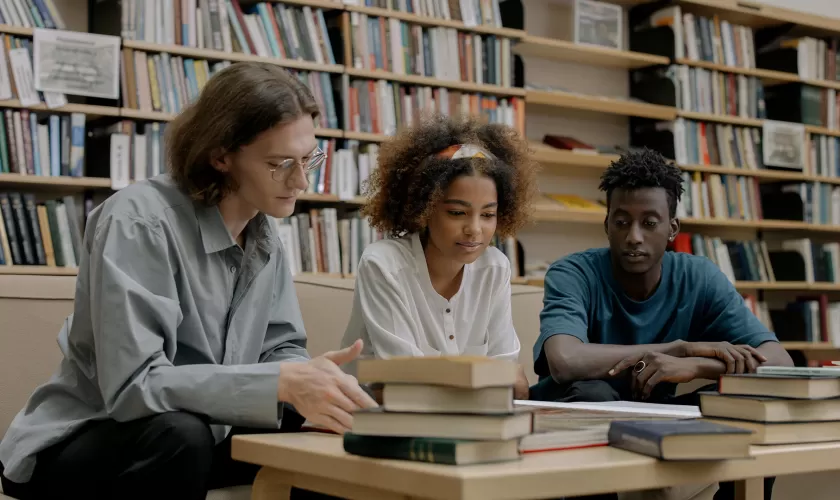 Drie studenten in een bibliotheek achter een tafel vol boeken. 2 mannen en in het midden een vrouw